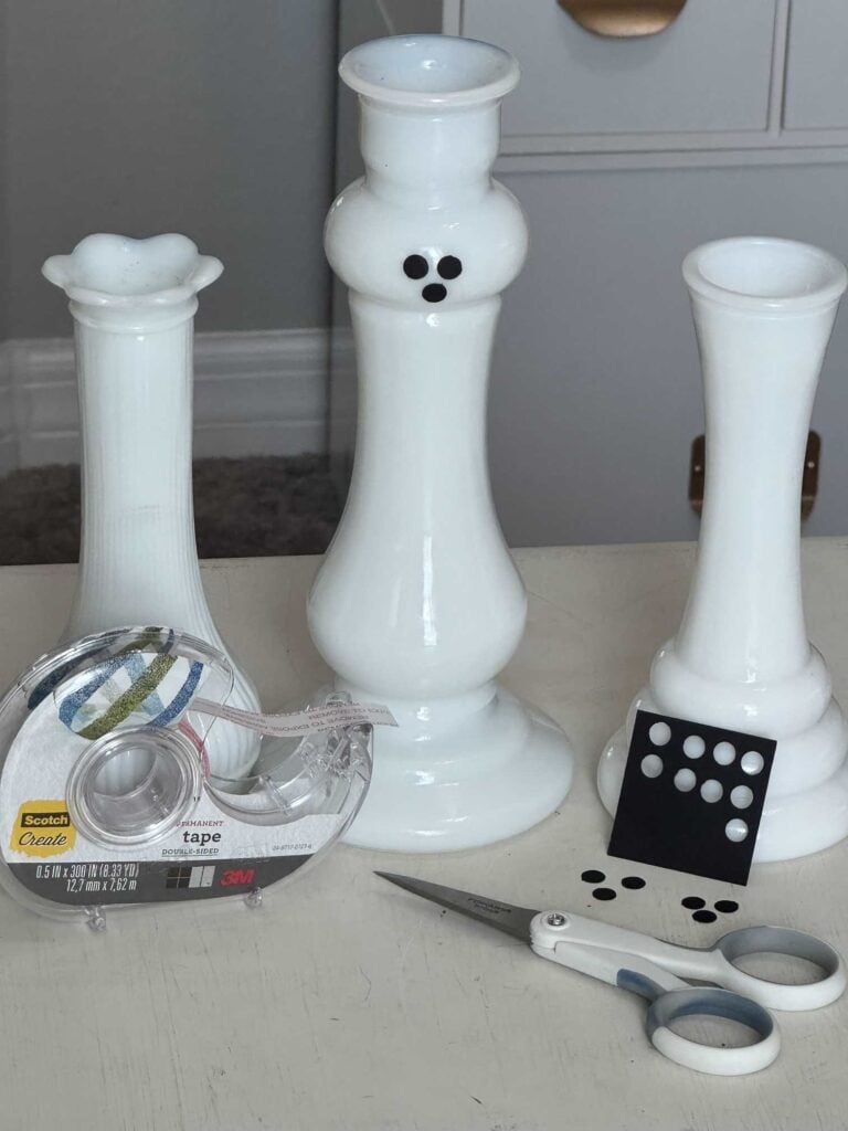 Three milk glass vases (one has eyes and a mouth) with black dots, a hole punch, scissors, and double-sided tape. 