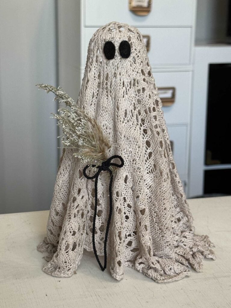 Ghost with black eyes made from an upcycled sweater and holding a small dried flower bouquet tied with black twine. 