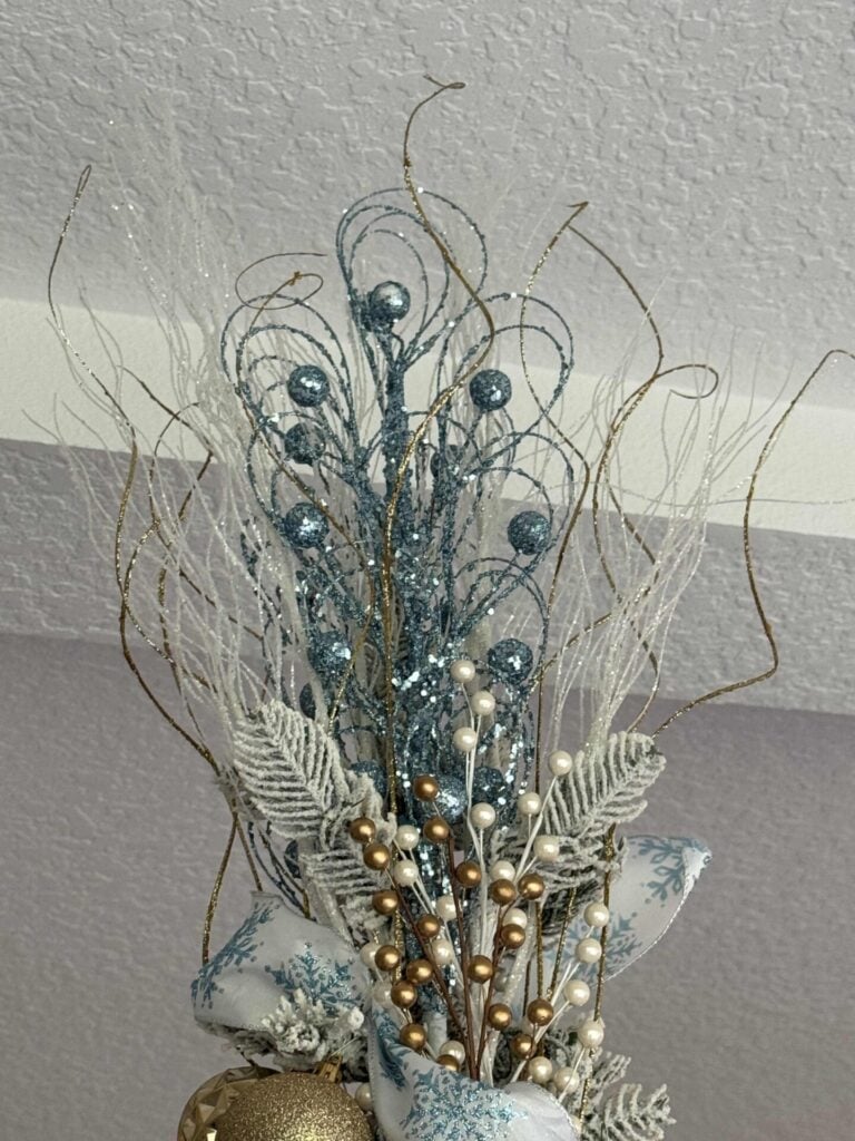 Step six to decorate a faux flocked Christmas tree: is to create a topper from blue, white, and gold sparkly picks.