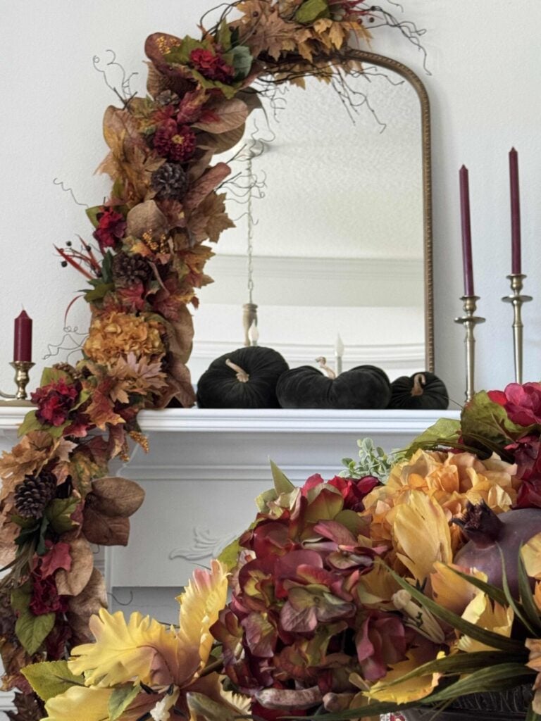 A fireplace mantel with green velvet pumpkins, three gold candlesticks with burgundy candles, and a fall garland draped over one side of a gold mirror.