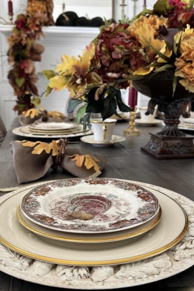 Brown transferware turkey plate stacked on ivory china and a leaf charger with matching centerpiece and garland in the background.