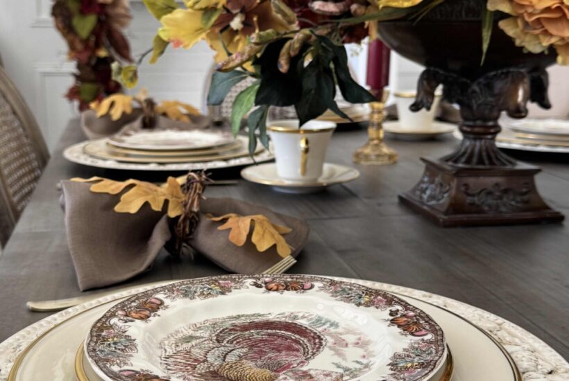 Brown transferware turkey plate stacked on ivory china and a leaf charger with matching centerpiece and garland in the background.