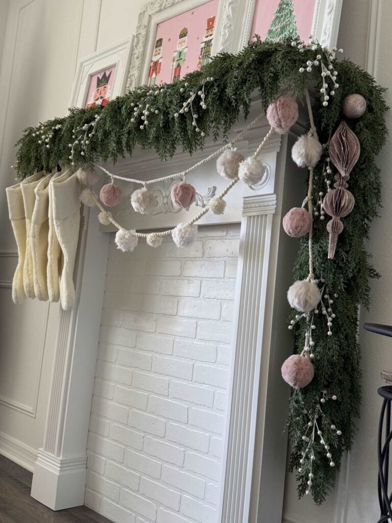 Mantel decorated with an evergreen garland, two pom pom garlands, stockings, white berries, and pink ornaments.