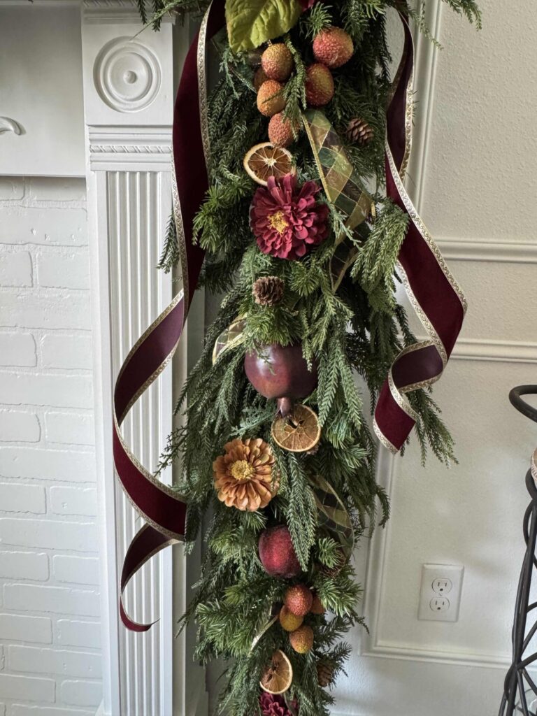 An evergreen garland decorated with faux flowers, fruit, burgundy ribbons, and dried orange slices that's hanging off a mantel. . 