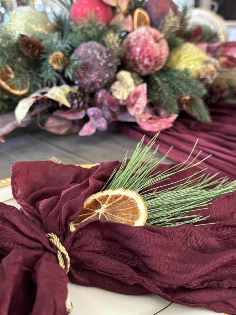 A burgundy napkin on a plate with an evergreen sprig and dried orange slice layered on it. 
