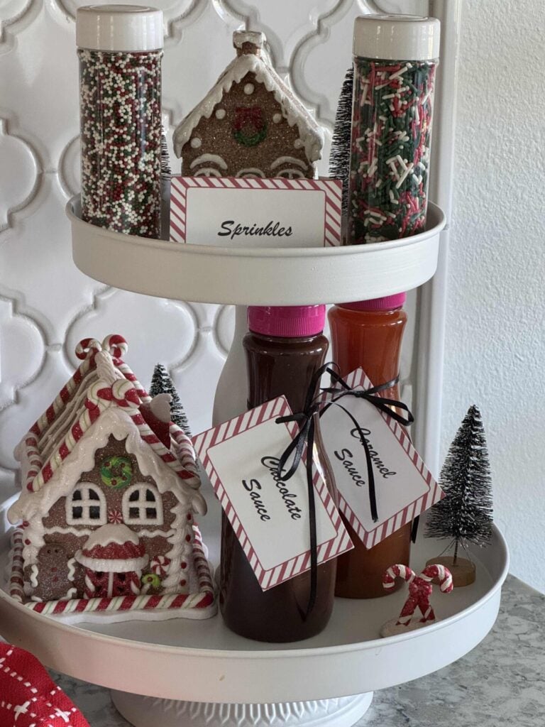 A white tiered tray holding sauces, sprinkles, and gingerbread decor. 