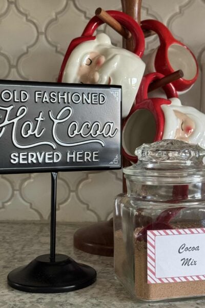 Sign that says "old fashioned cocoa served here sitting beside a jar of cocoa mix and Santa mugs.