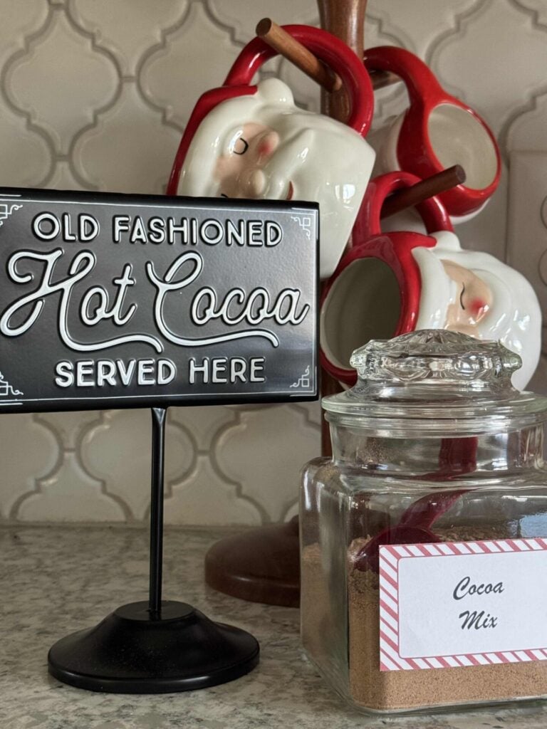 Sign that says, "old fashioned hot cocoa served here" next to a jar of hot chocolate mix and Santa mugs. 
