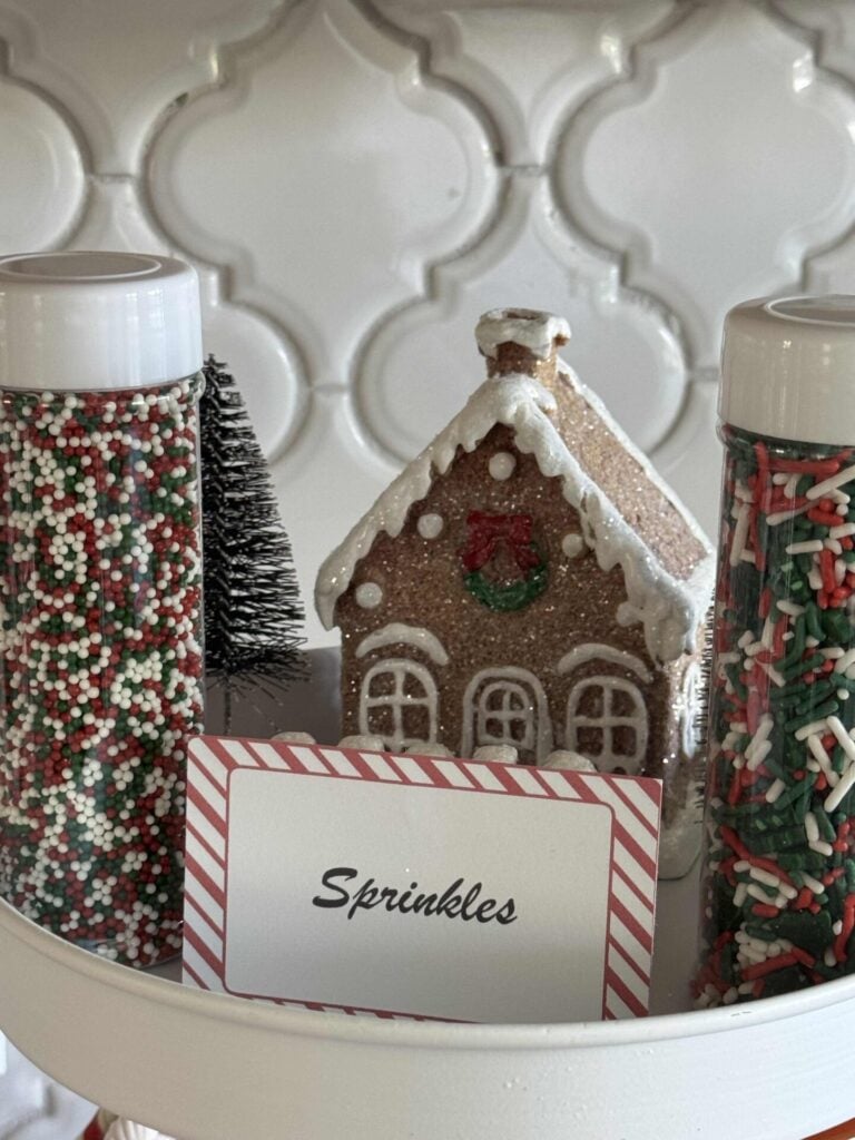 Two bottles of sprinkles on a tray with a label and faux gingerbread house. 