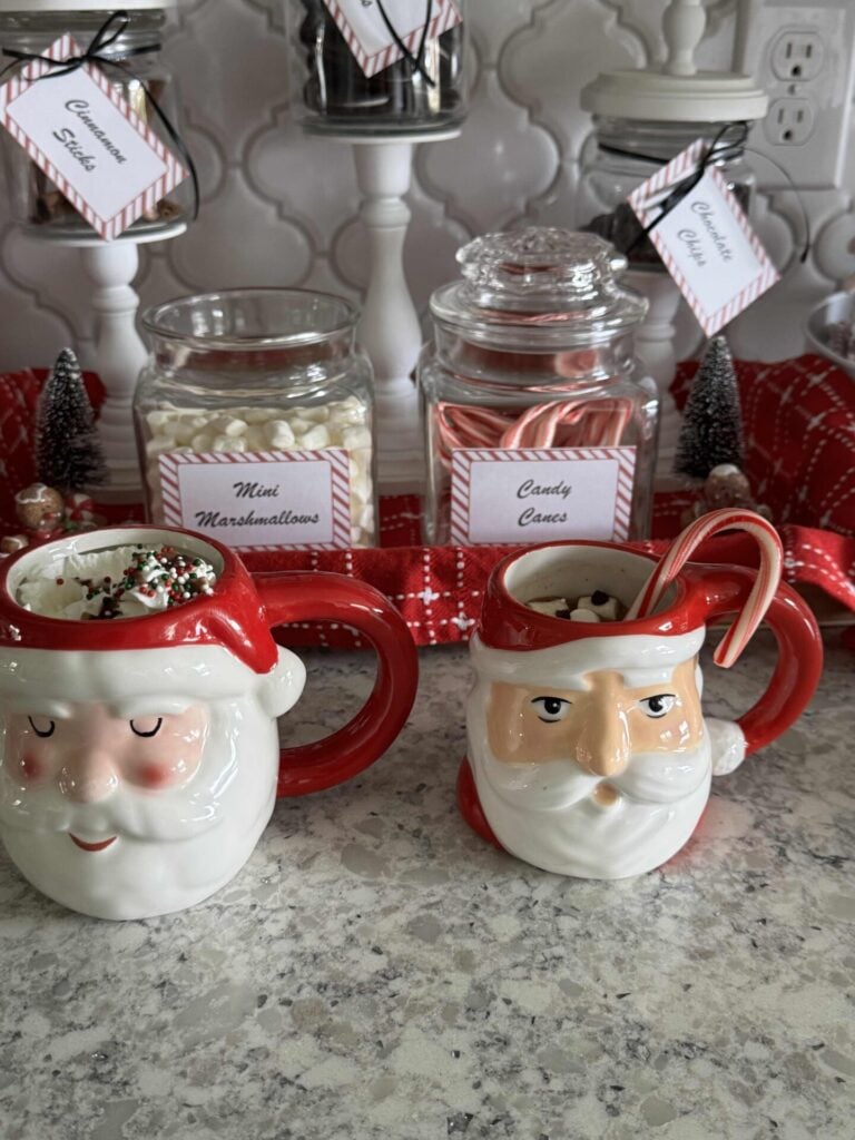 Two Santa mugs filled with hot cocoa and toppings in front of a hot chocolate bar. 