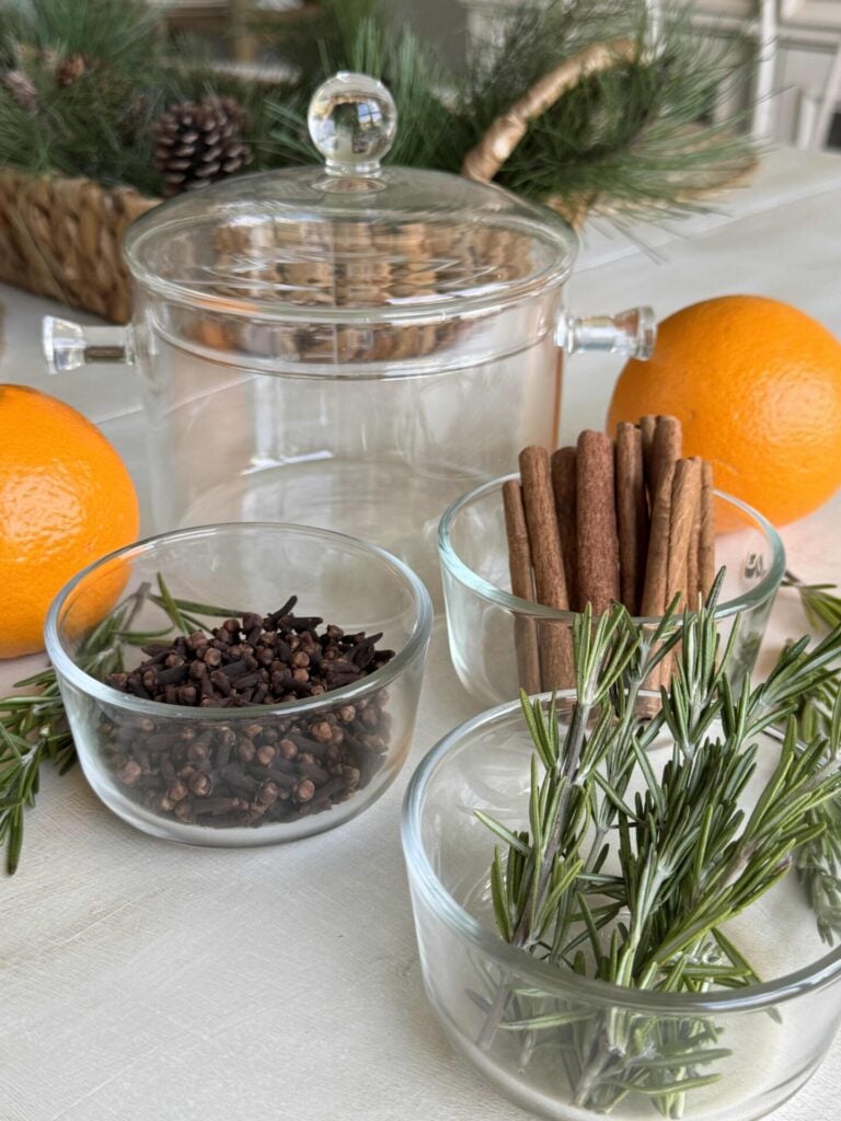 A glass simmer pot surrounded by oranges, and bowls of cloves, cinnamon sticks, and rosemary.
