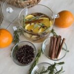 Winter simmer pot surrounded by oranges and bowls of cloves, cinnamon sticks, and rosemary.