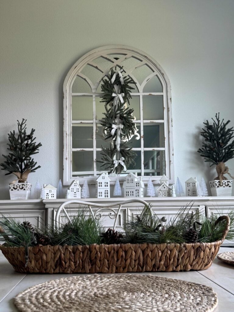 Basket on a table with buffet in background that holds white houses and tabletop trees with a triple mini wreath hanging on a mirror.