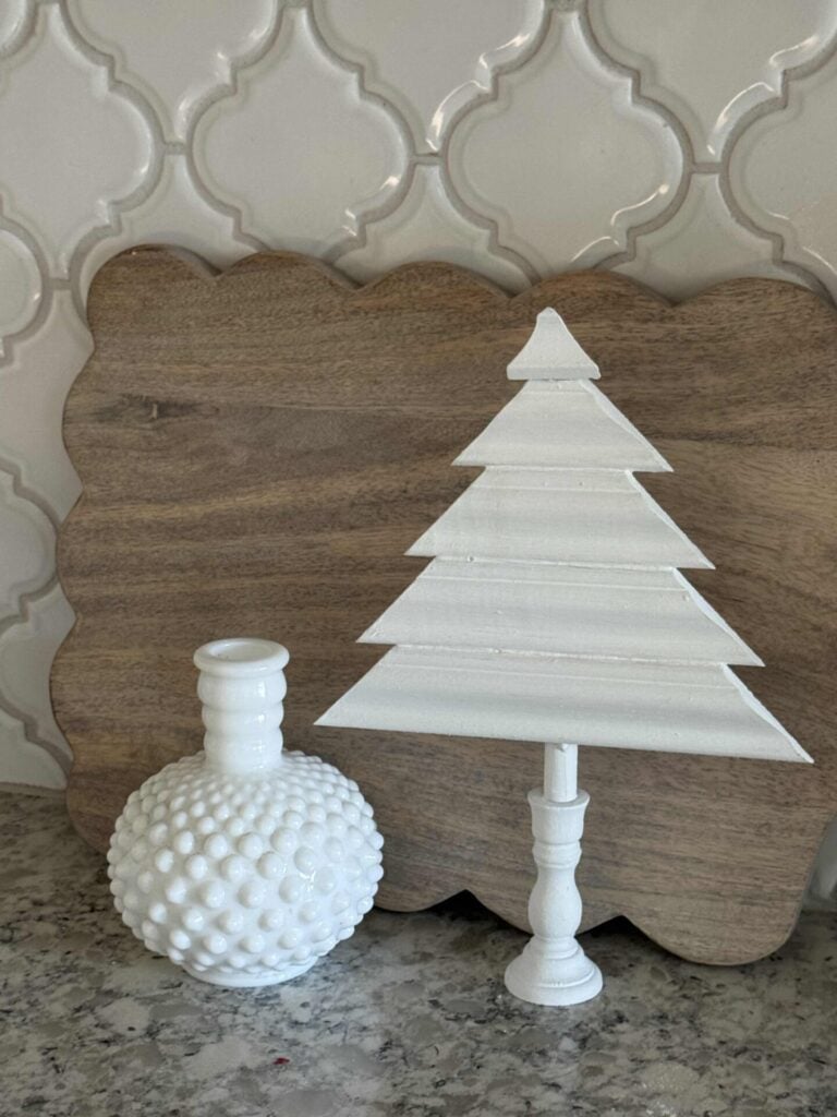 Cutting board leaning against a backsplash with a white wood tree and milk glass piece beside it. 