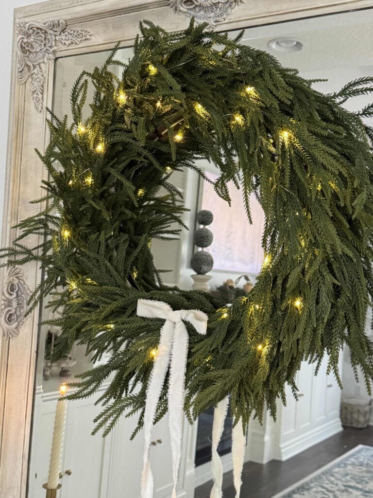 An evergreen wreath with fairy lights and white bow hanging on a mirror. 