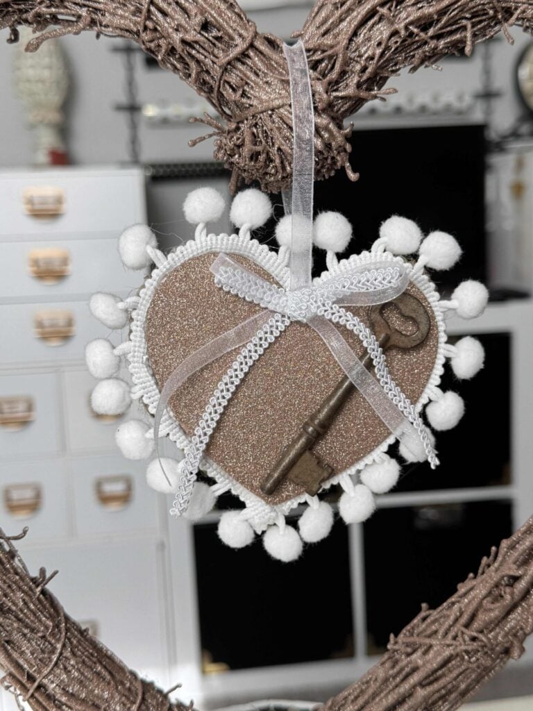 A glitter heart with white pom pom trim, a bow made with organza and gimp trim, and an antique key. 