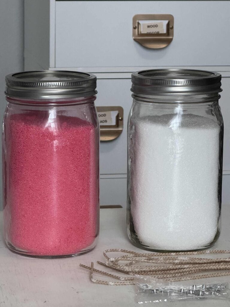 Two mason jars filled with sand candle wax, one is pink and the other white with a long wick and wick clips laying beside them.