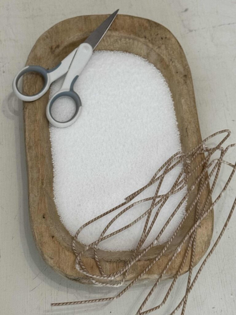 A wood bowl filled with white sand candle wax with a wick and scissors on it.