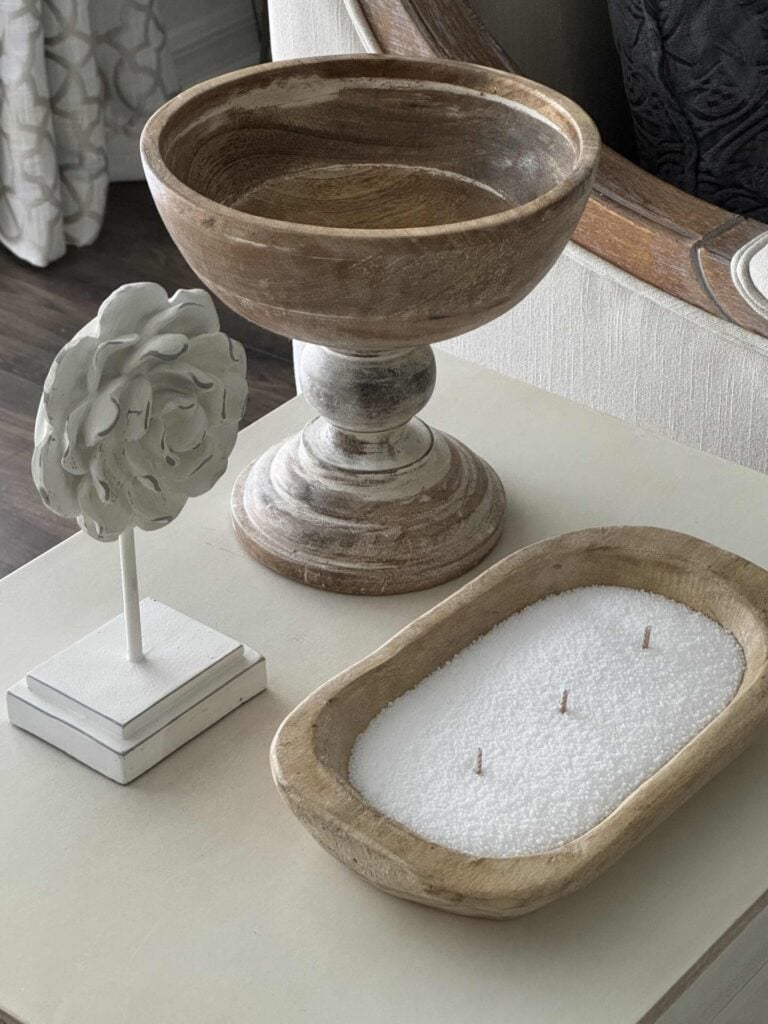 A DIY candle with three wicks in a wooden bowl next to a white flower statue and a wood pedestal bowl.