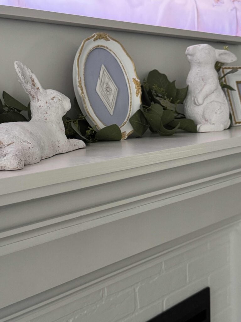 Two white ceramic bunnies, with blue and white art in between them,  on a mantel covered with a eucalyptus garland. 