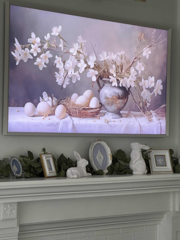 Frame TV with an image featuring soft dogwood branches arranged in a blue vase, paired with off-white eggs in a bowl, and scattered across the table. It's hanging over a living room mantel decorated with spring decor. 