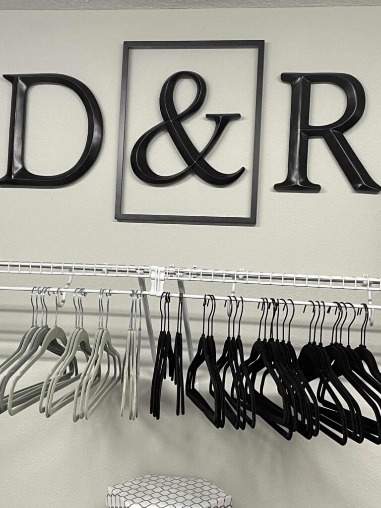 A bedroom closet with gray and black velvet hangers on a wire shelf. The letters D,R, and the & (inside a frame) symbol hang above it. 