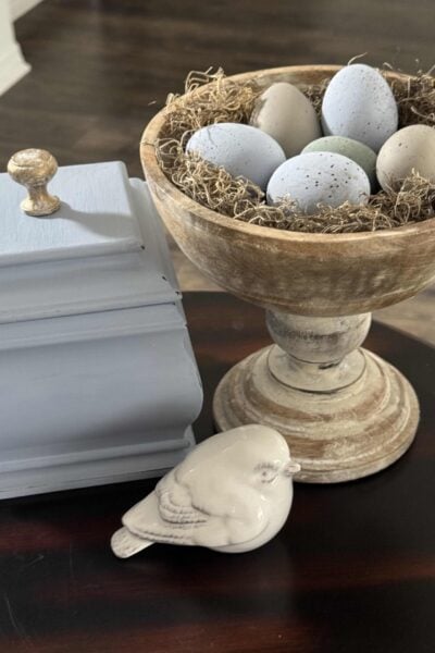 DIY speckled Easter eggs in a wood bowl, next to a blue box and a white ceramic bird.