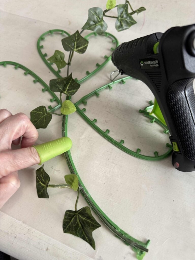 Someone hot gluing ivy to a shamrock form while wearing finger protectors. 