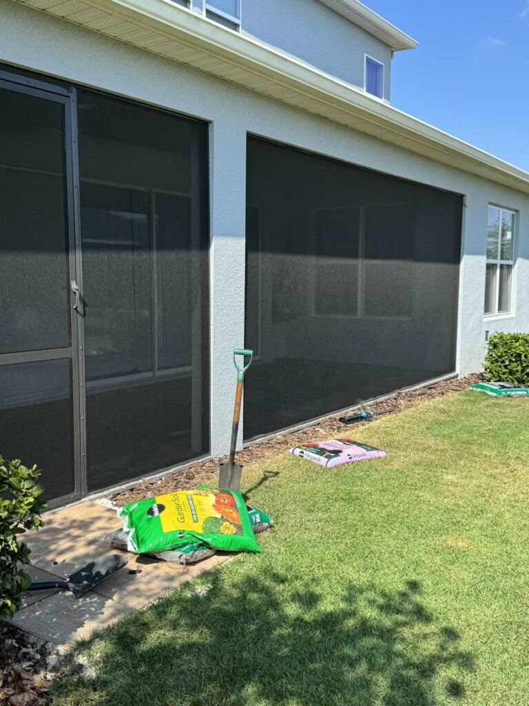 A screened in porch and yard with bags of soil , mulch, and a shovel. 