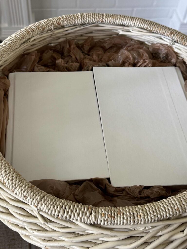 A white vintage basket with books stacked inside it and plastic grocery bags tucked around the books. 