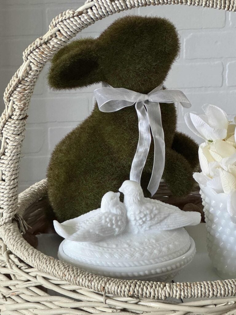 A white vintage basket with a moss bunny, milk glass bowl with tulips, and a milk glass lovebird trinket dish inside.