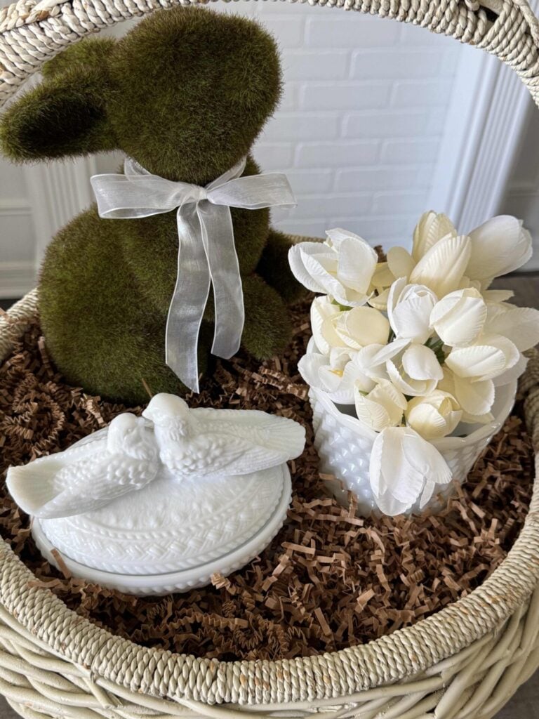 A white vintage basket with a moss bunny, milk glass bowl with tulips, and a milk glass lovebird trinket dish surrounded by brown crinkle paper. 
