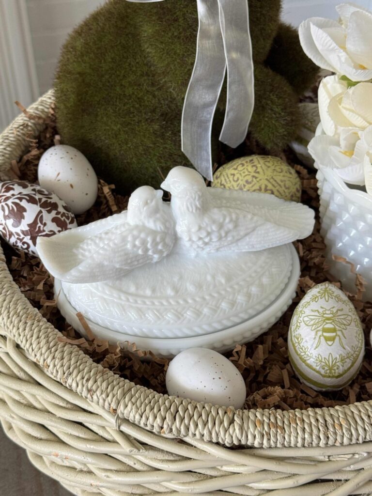 A white vintage basket with a moss bunny, milk glass bowl with tulips, and a milk glass lovebird trinket dish surrounded by brown crinkle paper with decoupage eggs scattered around. 