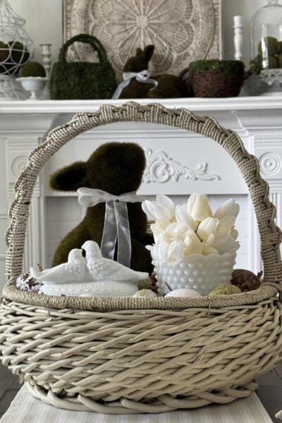DIY Easter Centerpiece in a white basket with a moss bunny, milk glass bowl with tulips, and a milk glass lovebird trinket dish, surrounded by decoupage eggs.
