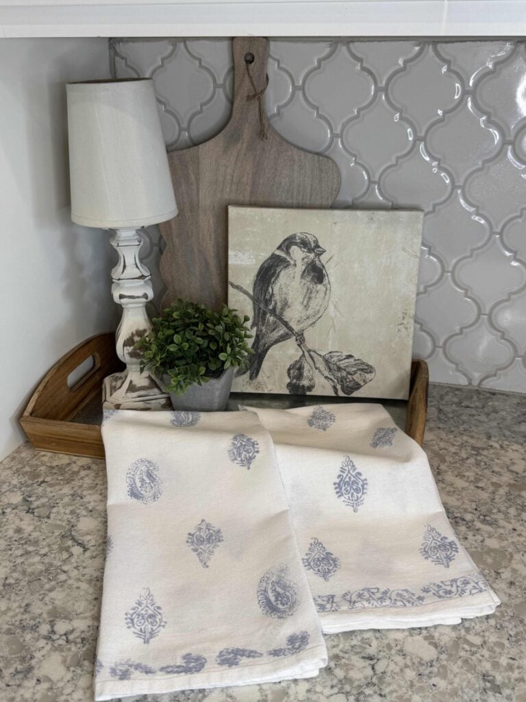 Spring kitchen decor idea #5 is to create a vignette with a tray, propped up wood cutting board, a spring bird canvas, a cordless lamp, a small faux herb, and block print kitchen towels draped over the tray. 