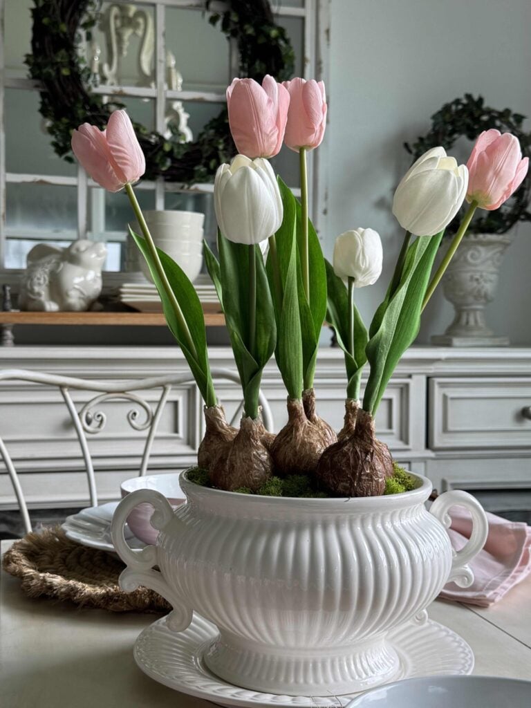 DIY tulip bulb arrangement in a white soup tureen sitting on a matching plate, on a kitchen table. 