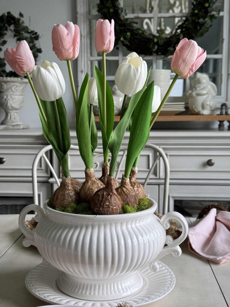 A DIY tulip bulb arrangement in a white soup tureen sitting on a matching plate, on a kitchen table. 