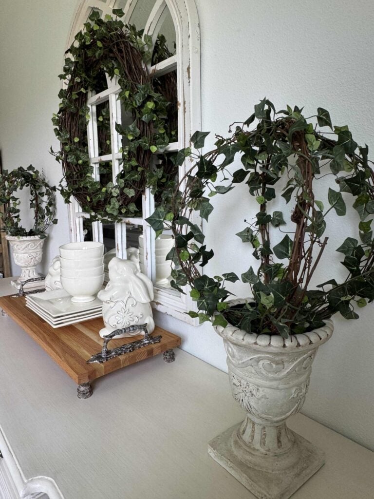 A grapevine wreath and two topiaries covered in ivy decorating a buffet. A tray with off white dishes and bunnies sit on the buffet. 