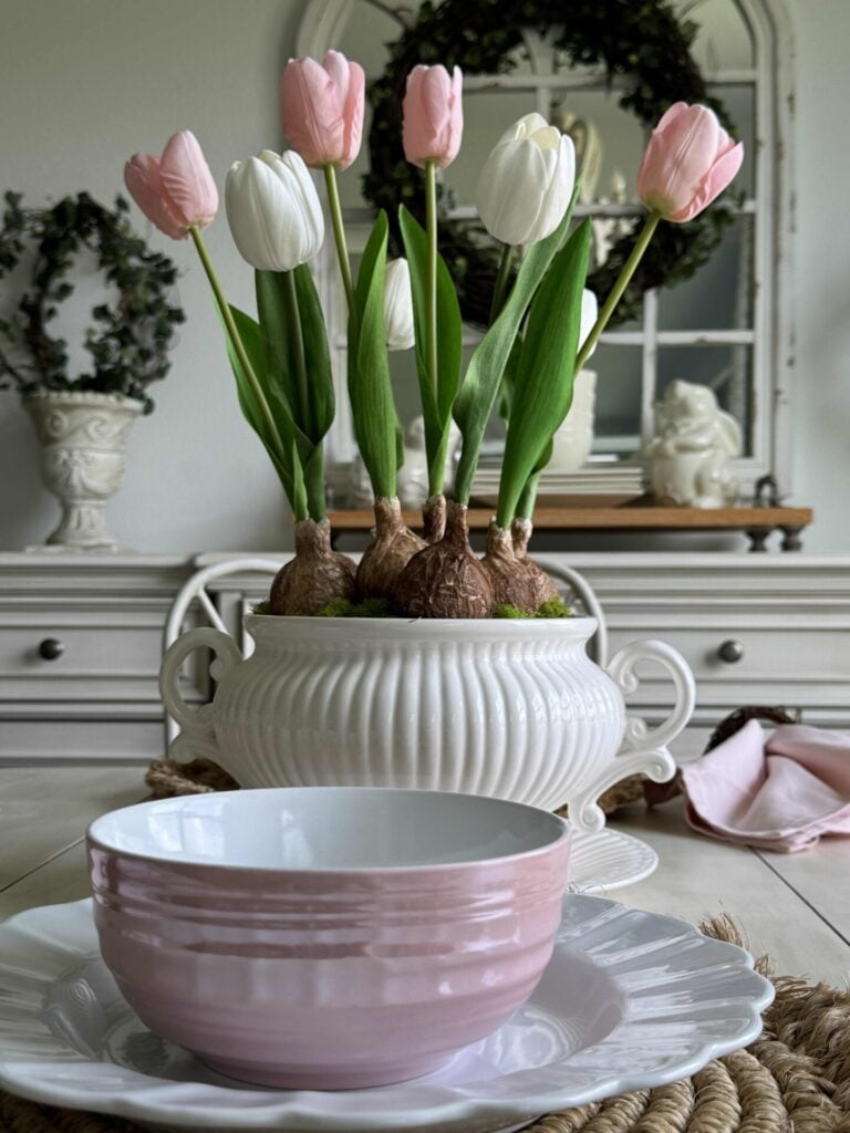 A kitchen table decorated with white plates, pink bowls, and a pink and white tulip bulb arrangement. 