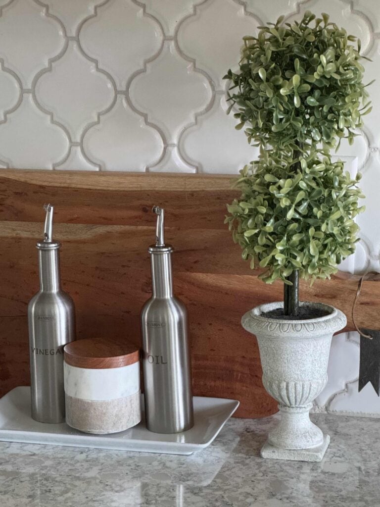A vignette with a wood cutting board propped up with a white tray in front of it holding a salt and pepper container and two vessels with oil and vinegar. Next to it is a topiary. 