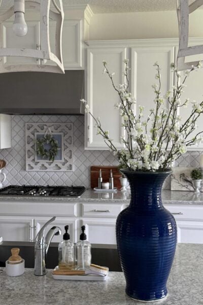 A tall blue vase filled with flowering branches sits on a kitchen counter.