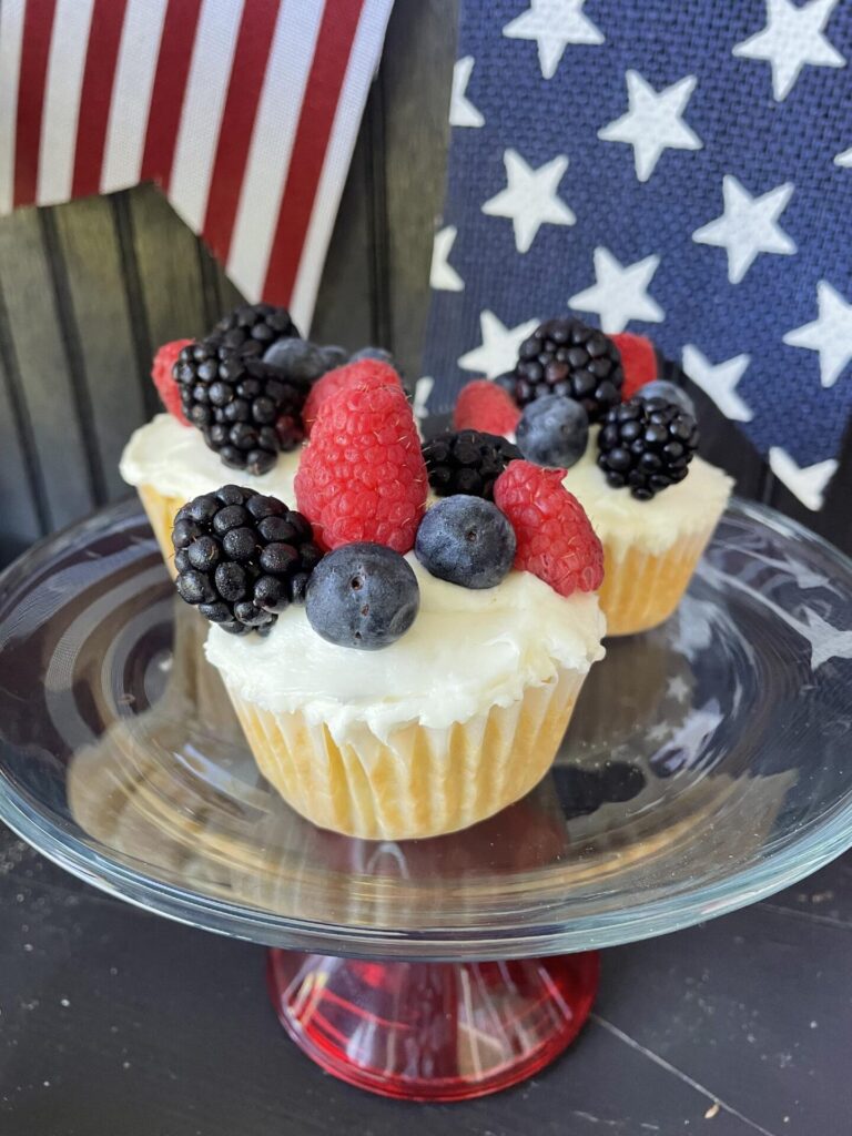Cupcakes topped with white frosting, blackberries, raspberries, and blueberries are displayed on a glass cake stand—perfect for patriotic entertaining ideas. In the background are American flag-themed decorations with stars and stripes.