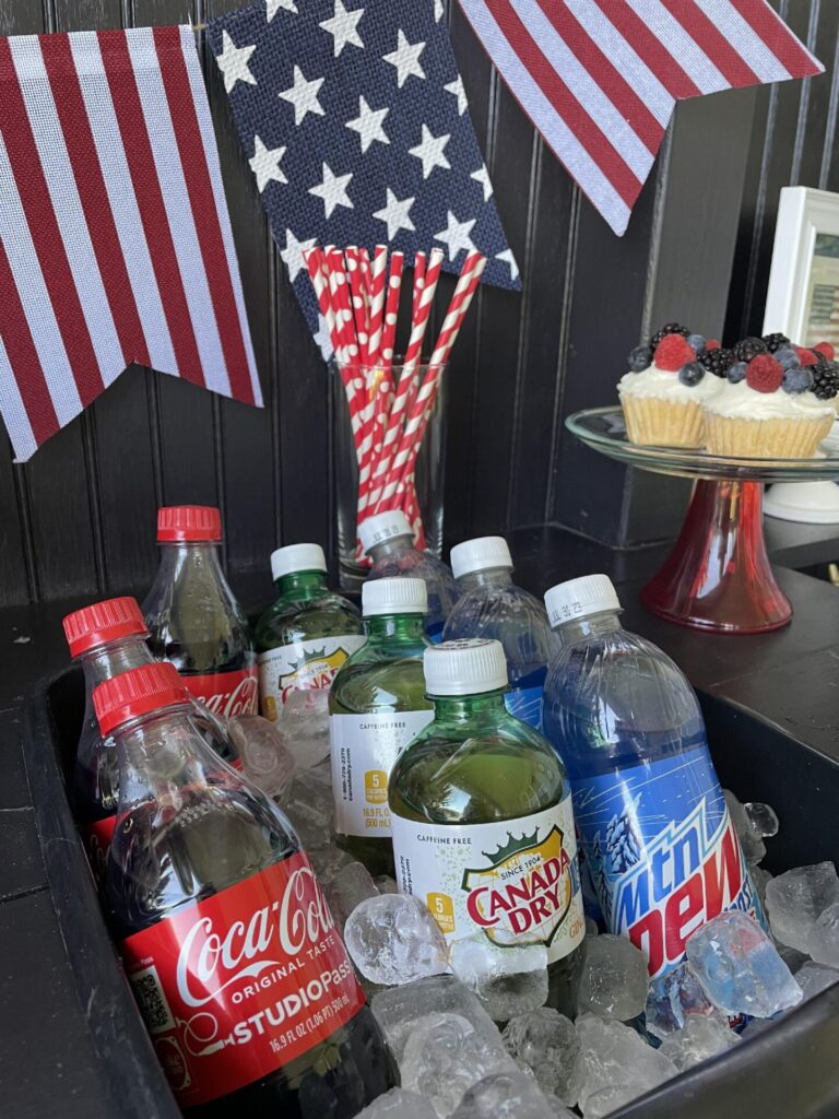 Bottles of Coca-Cola, Canada Dry, and Mountain Dew are chilled in ice, with American flag bunting, red-and-white striped straws, and a berry-topped cupcake—perfect for Patriotic Entertaining Ideas—on a cake stand in the background.