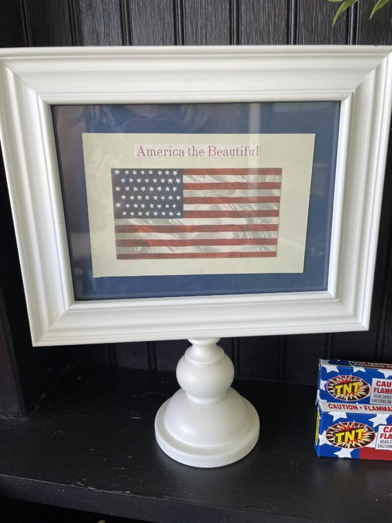 A framed print of an American flag with "America the Beautiful" sits on a white pedestal stand, perfect for Patriotic Entertaining Ideas. Two small boxes of TNT fireworks rest beside it against a dark wood background.