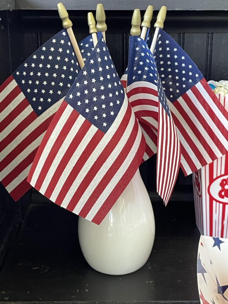 A white vase holds several small American flags with red, white, and blue colors—perfect for Patriotic Entertaining Ideas. Nearby, a container with a red and white striped popcorn design stands out against the dark background.