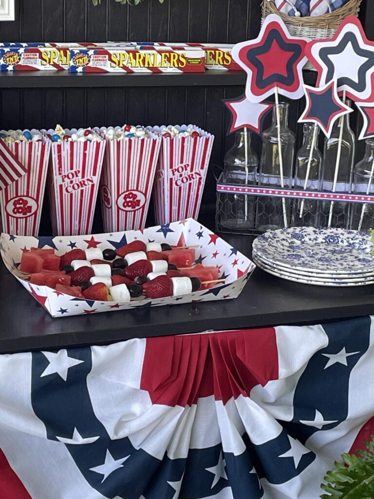 A festive table with popcorn in striped containers, fruit skewers of strawberries, marshmallows, and watermelon, blue and white plates, and star decorations—perfect for patriotic entertaining ideas with a red, white, and blue theme.