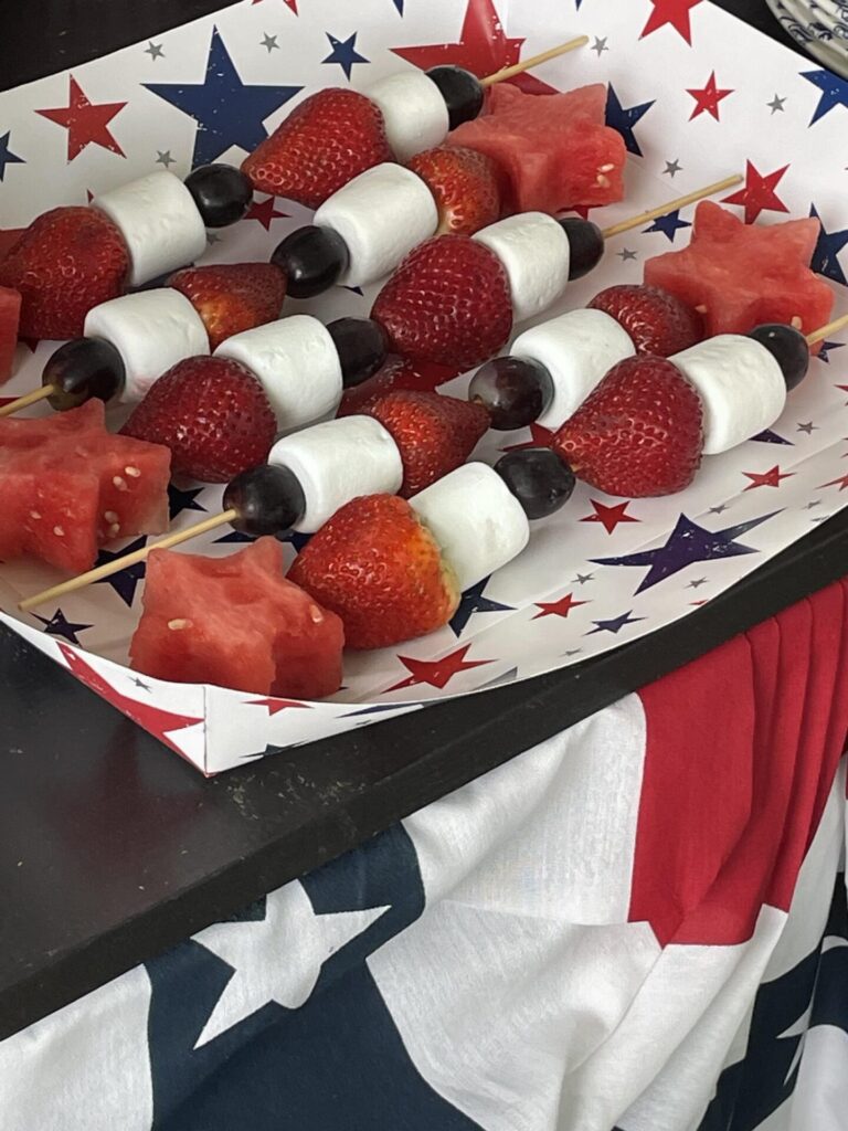 Skewers with strawberries, grapes, marshmallows, and star-shaped watermelon pieces are arranged on a star-patterned tray, perfect for patriotic entertaining ideas on a red, white, and blue tablecloth.