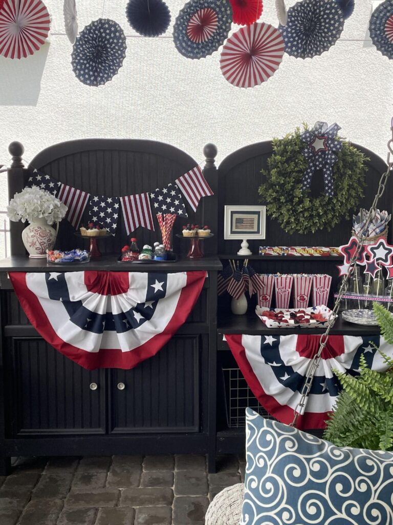 Patriotic-themed party setup with red, white, and blue decorations, American flags, bunting, treats, popcorn, a green wreath on a black cabinet outdoors, and festive fan decor above—perfect for inspiring Patriotic Entertaining Ideas.