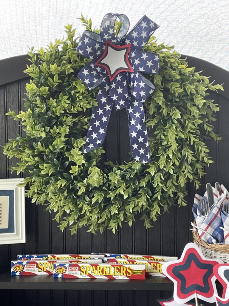 A green wreath with a blue and white star-patterned bow and red-bordered star hangs on a dark wall. Below, find boxes labeled Sparklers—perfect for Patriotic Entertaining Ideas—and star-shaped decorations with napkins and utensils nearby.