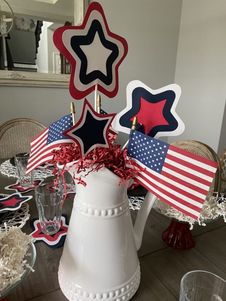 A white pitcher holds red, white, and blue star decorations and small American flags with red shredded paper inside—a festive touch perfect for Patriotic Entertaining Ideas on a dining table set with glasses and star-shaped coasters.